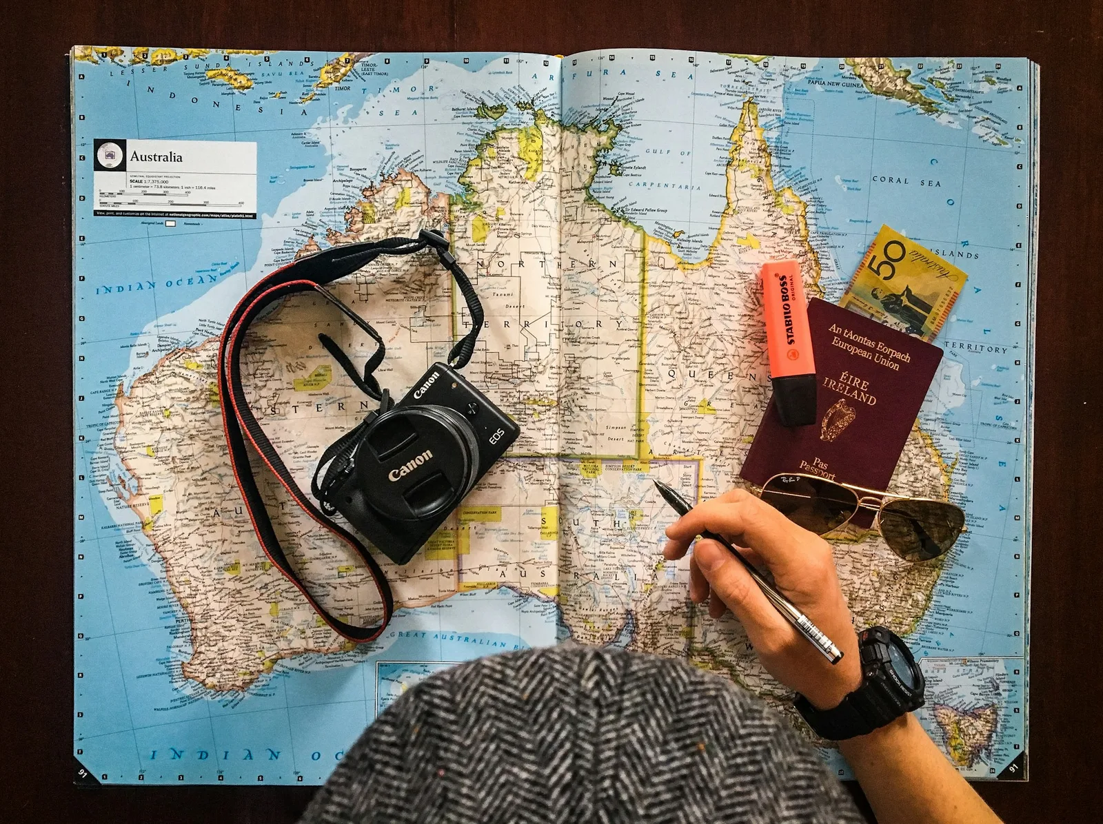 A hand holding a pen over a map of Australia beside a camera and passport.