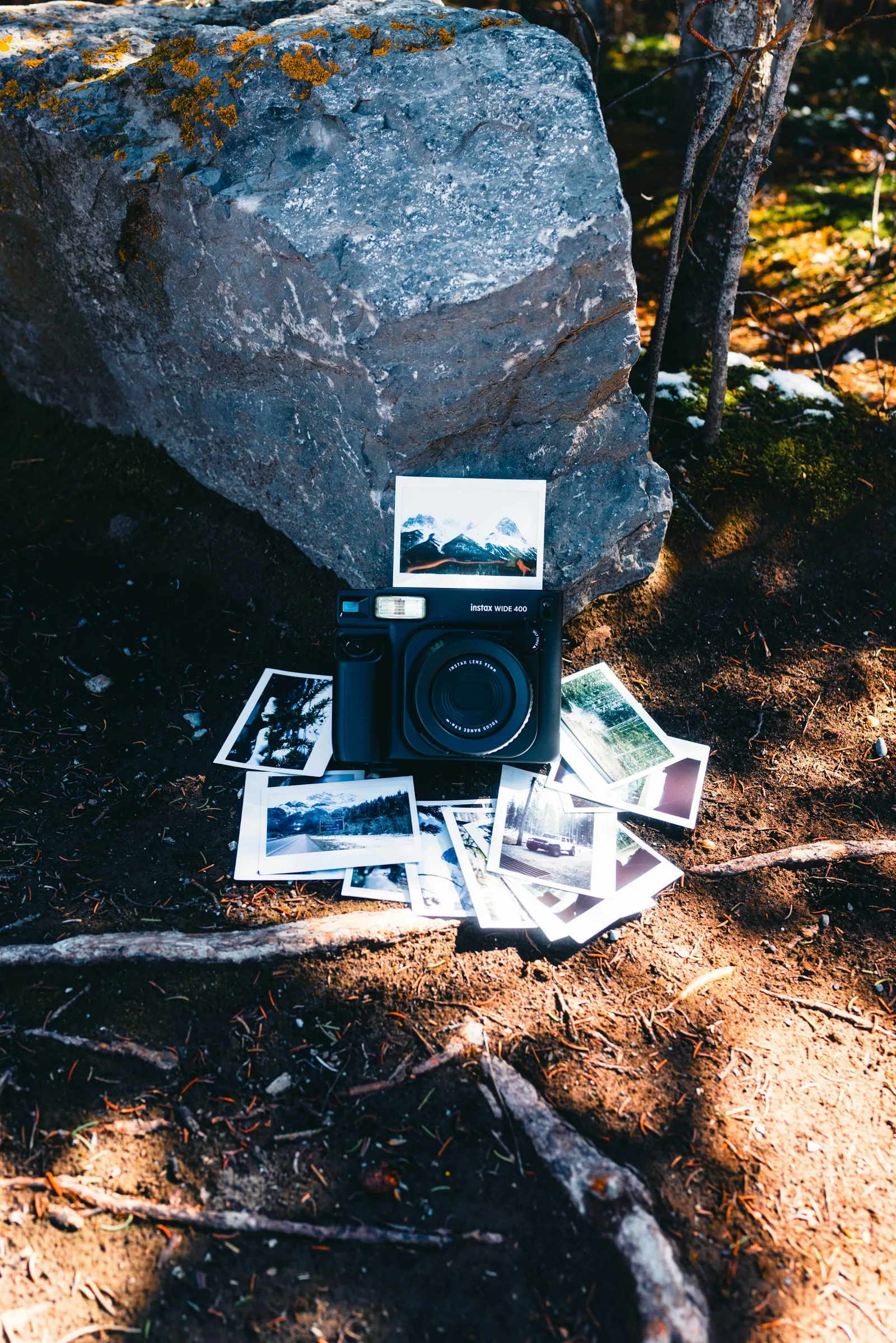 An instant camera and printed landscape photographs beside a rock in a forest.